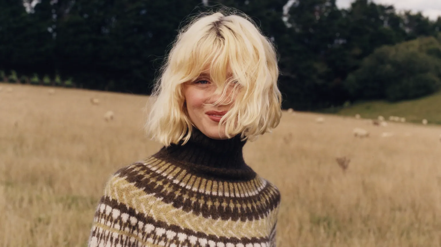 Ella Pavlides Maisie Peters wears a high neck brown and yellow knit jumper and has her blonde hair cut into a wavy bob. She stands against a field with crops and sheep