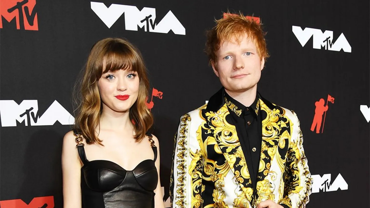 Getty Images Maisie Peters and Ed Sheeran on the red carpet at an MTV event. A young Maisie Peters has brown hair and wears black ankle boots with heels and a black leather dress with buckle straps. Ed Sheeran wears a yellow paisley patterned blazer with black trousers and shoes and a black shirt underneath